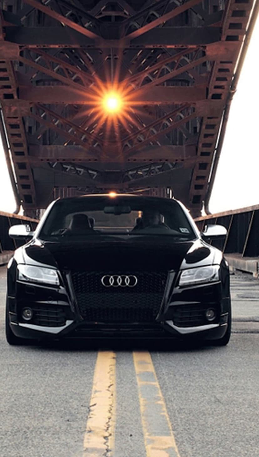 Black Audi sedan on an urban bridge with sunbeams filtering through a metal truss structure during sunset.
