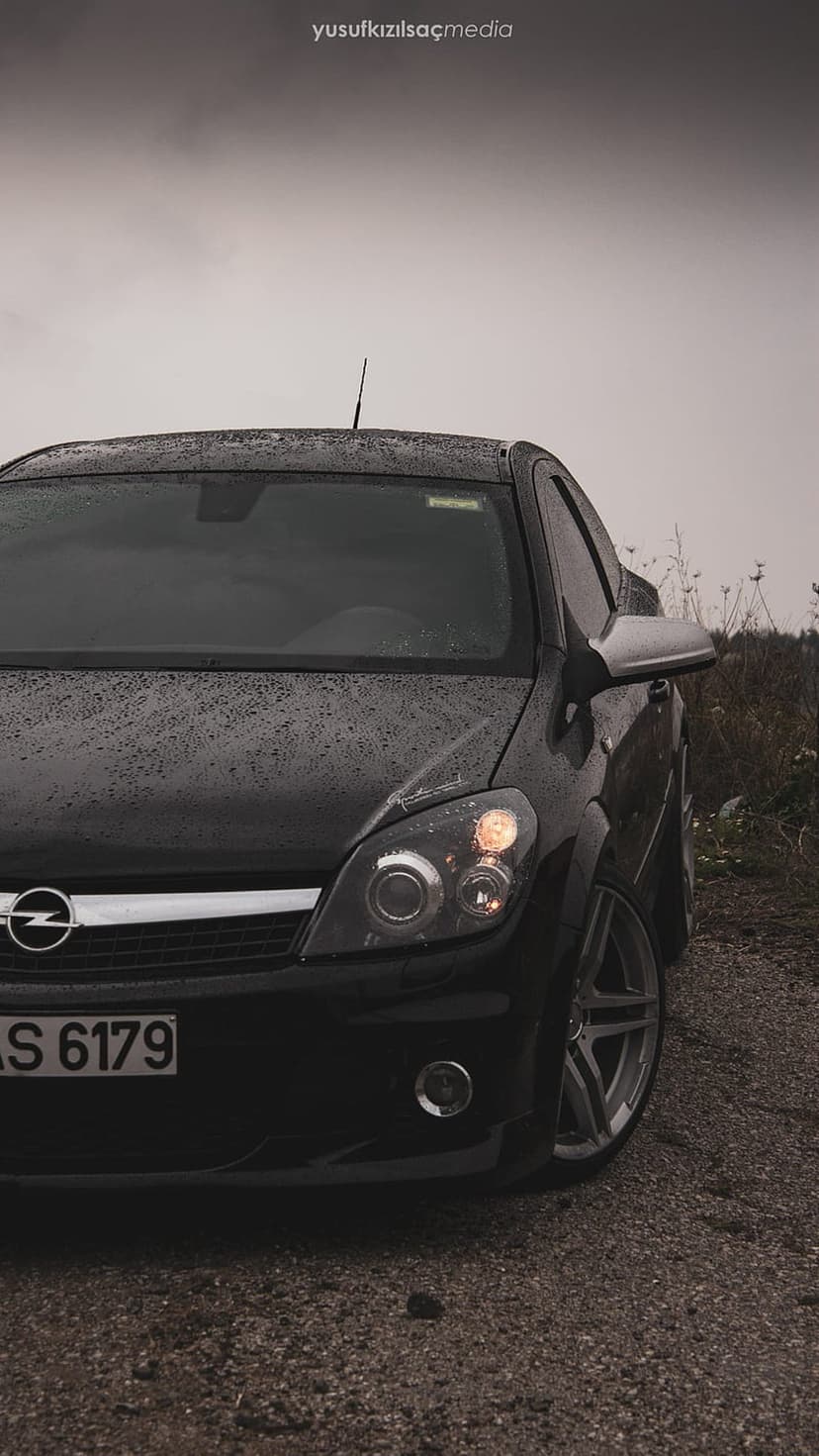 Black Opel Astra coupe parked on wet gravel with illuminated headlights under a cloudy, overcast sky