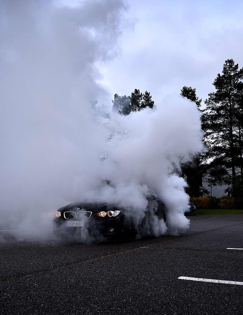 Black BMW sports car generates a thick cloud of white tire smoke on asphalt with pine trees in the background.