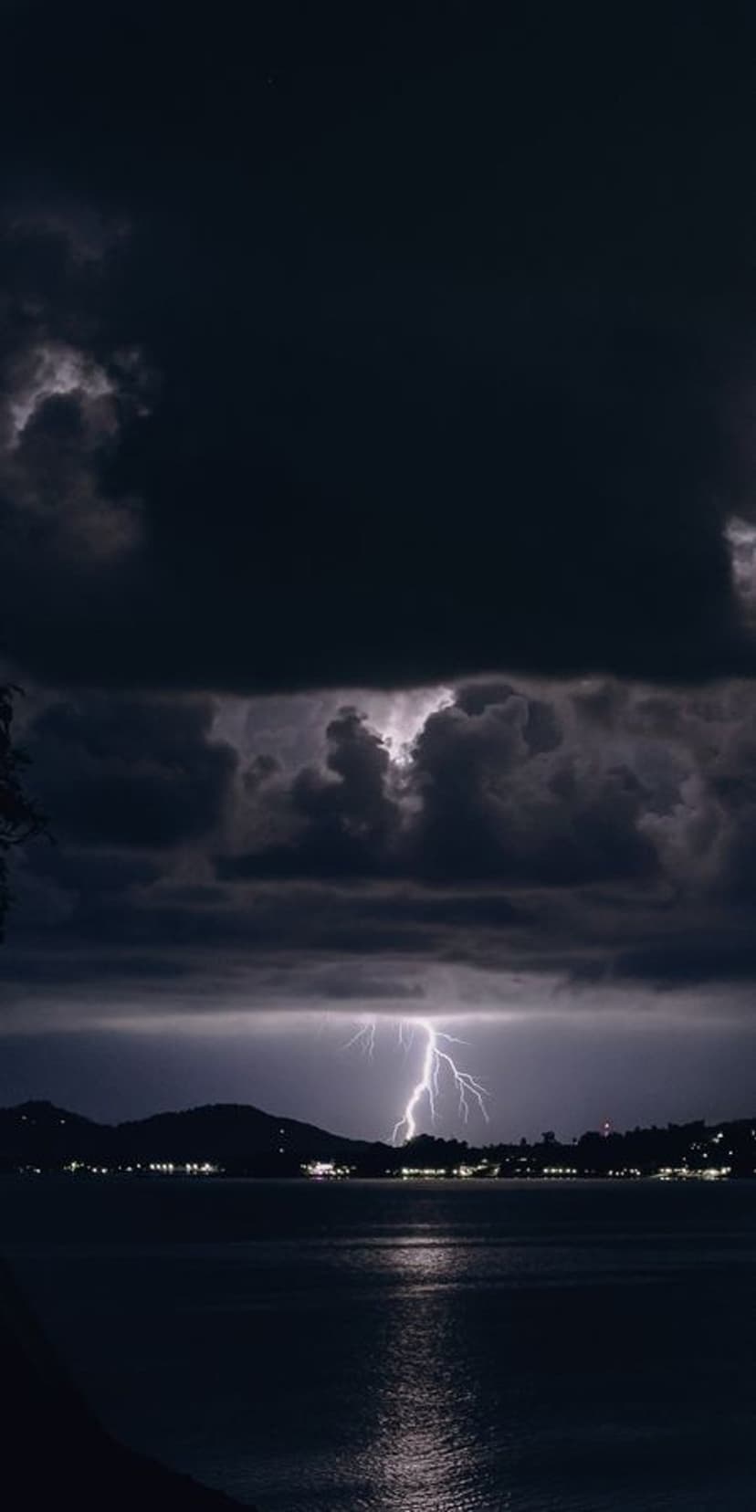 A jagged white lightning bolt strikes a dark body of water with silhouetted mountains and city lights at night.