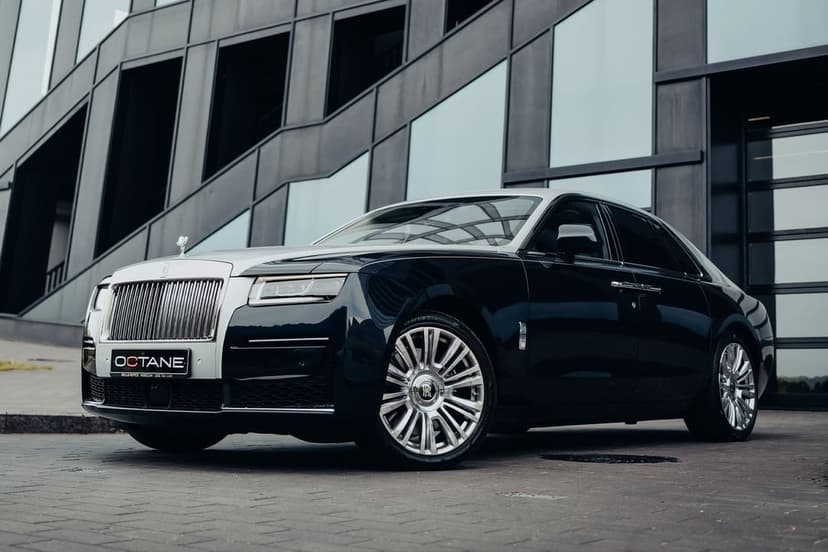 Two-tone Rolls-Royce Ghost parked in front of dark geometric modern architecture on a stone pavement.