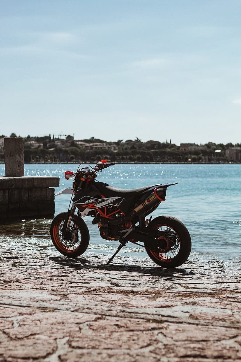 KTM supermoto motorcycle in orange and black parked on a cobblestone path beside a calm blue waterfront scene.