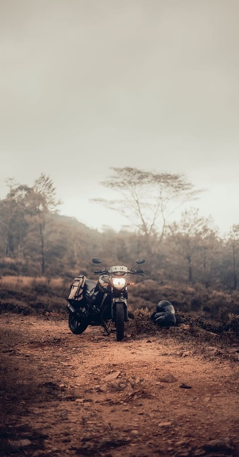 Vintage motorcycle parked on a gravel path within a dense foggy forest with a helmet resting on the ground nearby.