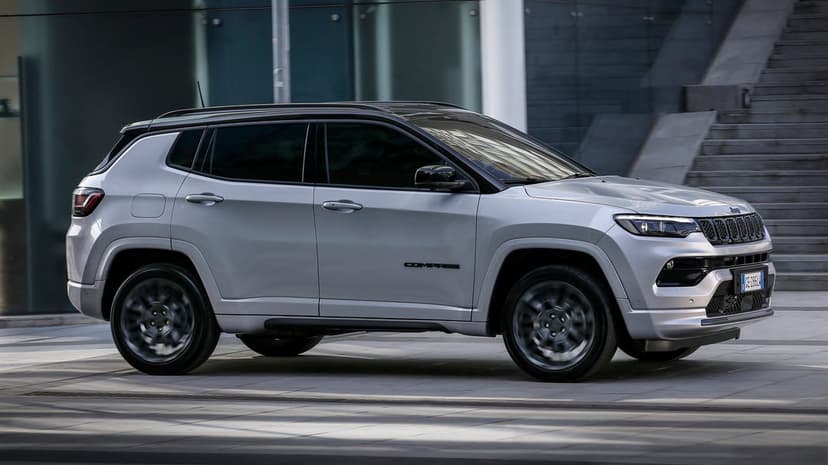 Silver Jeep Compass driving past a modern glass building with motion-blurred wheels in bright daylight.