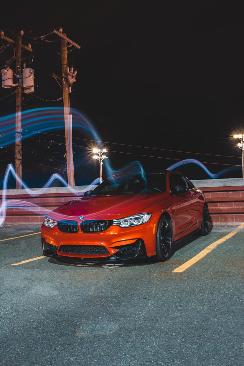 Orange BMW M4 parked at night with swirling blue and red light trails creating a long exposure motion effect.