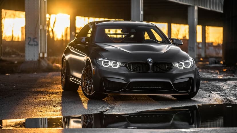 Matte black BMW M4 GTS at sunset with glowing headlights reflected in a puddle under a bridge.