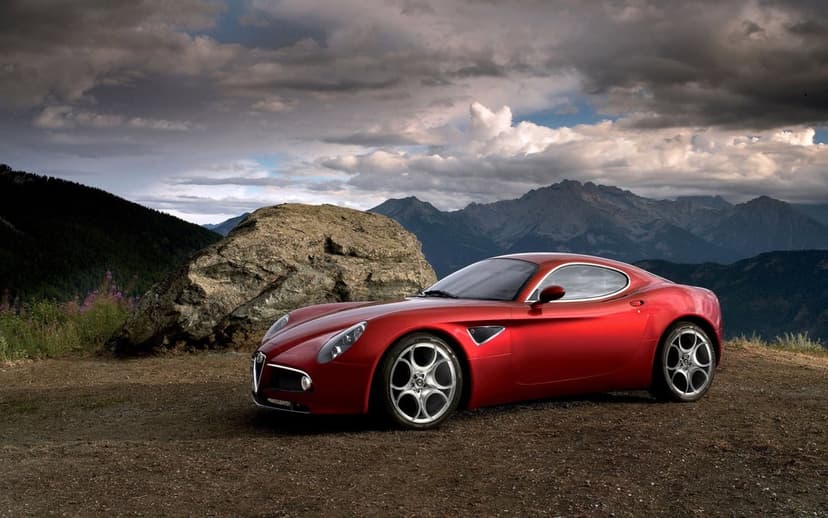 Red Alfa Romeo sports car on a dirt road with large rocks and distant mountains under a cloudy sky.