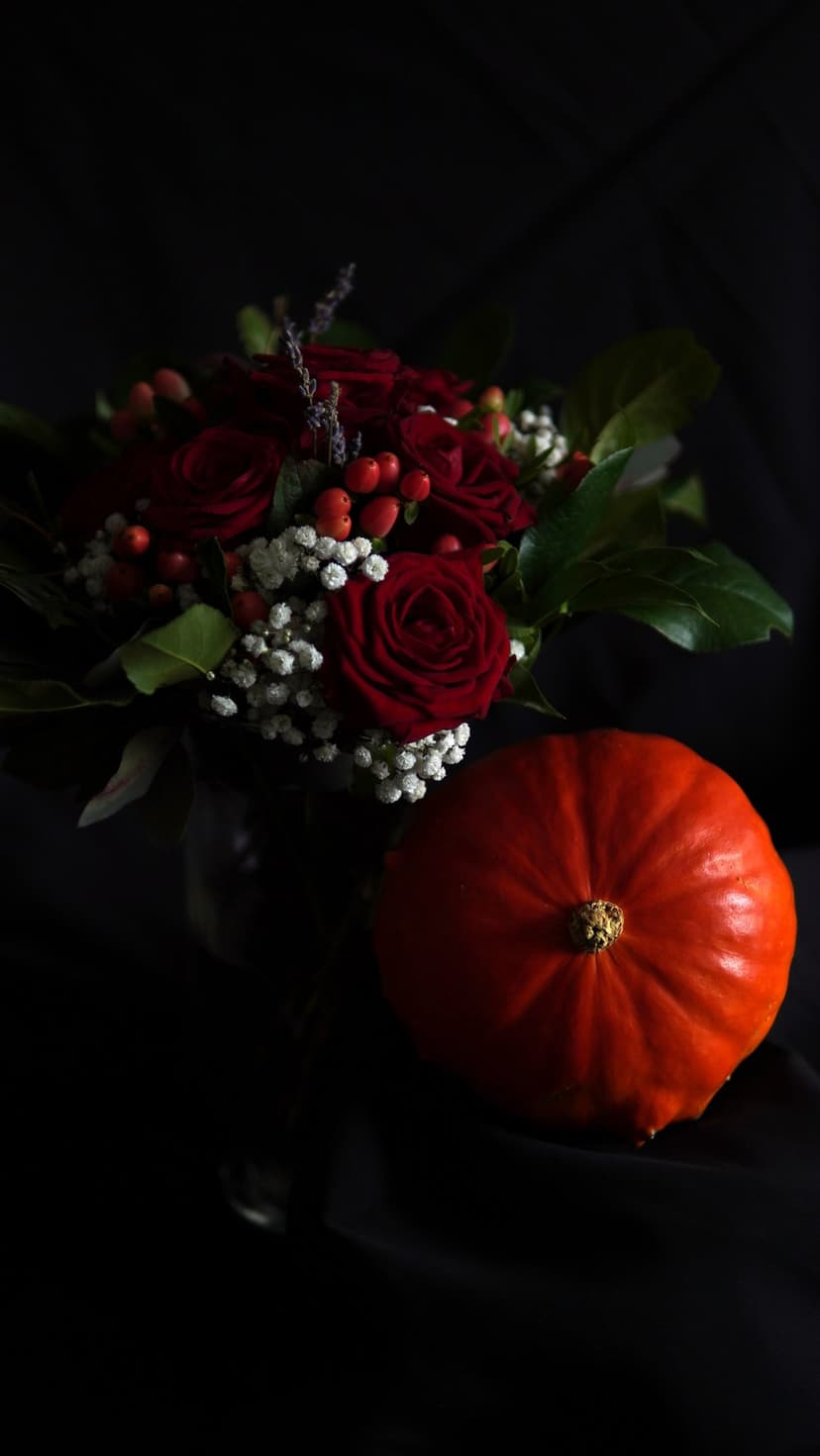 Deep red roses and a bright orange pumpkin sit against a dark textured backdrop with small white flowers.