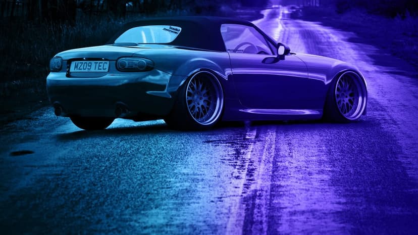 Stanced Mazda MX-5 NC convertible on a road with deep-dish wheels and vibrant blue and purple neon lighting.