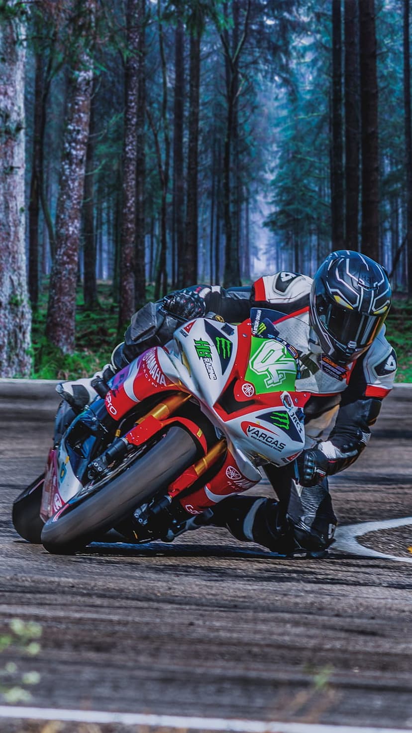 Motorcycle racer in a black suit leans low on a track surrounded by misty trees and dense forest foliage.