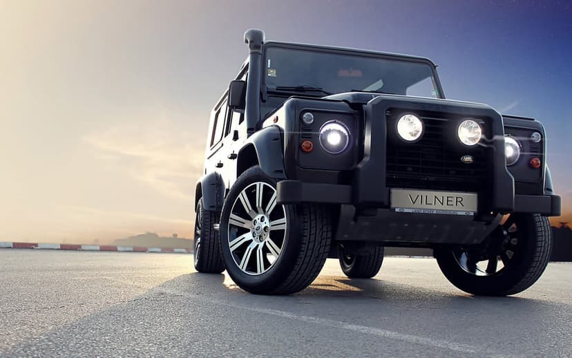 Black Land Rover Defender Vilner with glowing headlights parked on asphalt during a vibrant golden sunset.