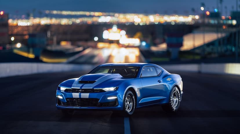 Blue Chevrolet COPO Camaro with silver stripes on a night track featuring soft bokeh city lights background.