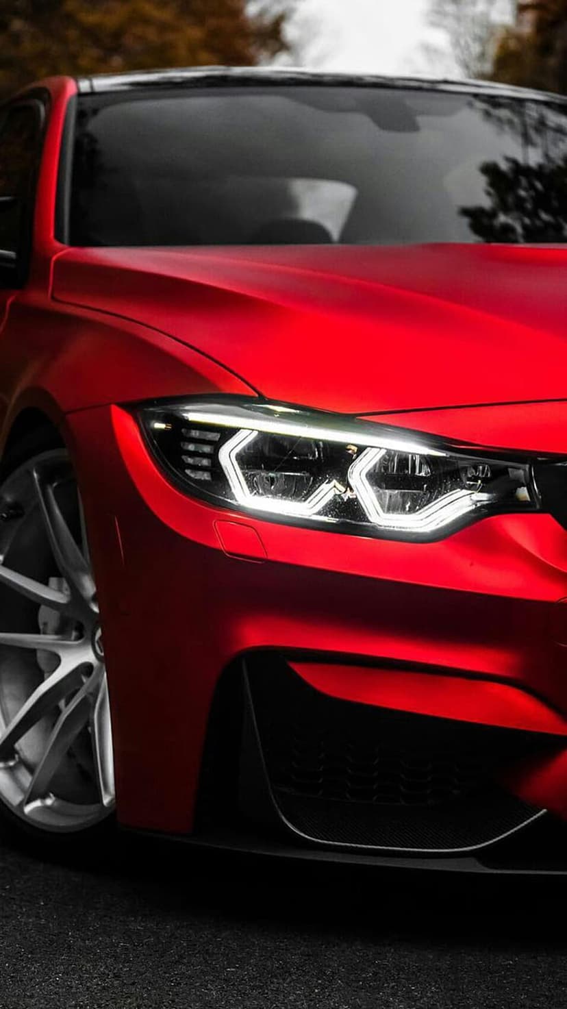 Red BMW M4 sports car features sharp LED headlights and dark multi-spoke wheels against a blurred green backdrop.