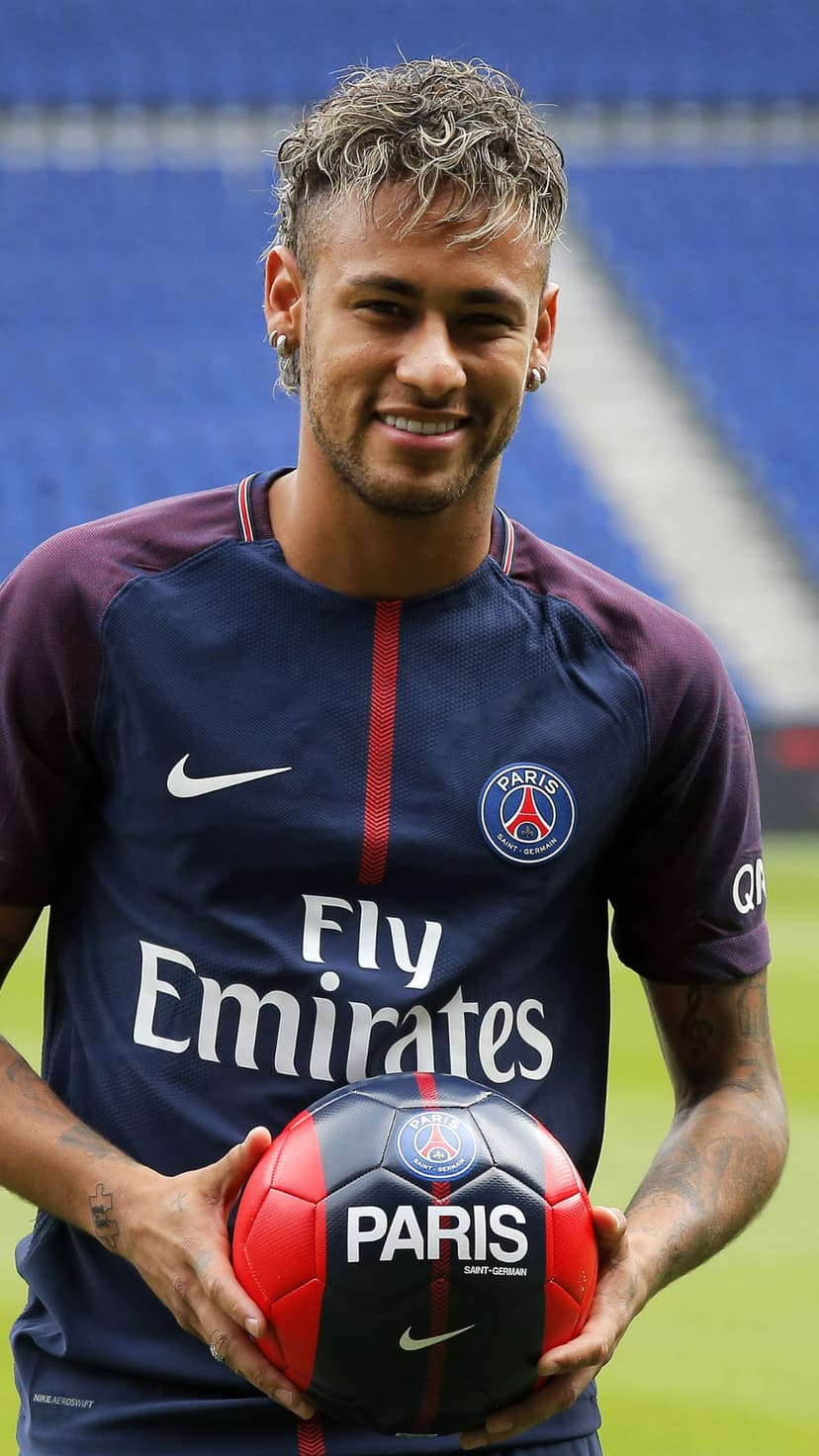 Neymar Jr. smiles in a navy PSG jersey while holding a soccer ball against blue stadium seats in the background.