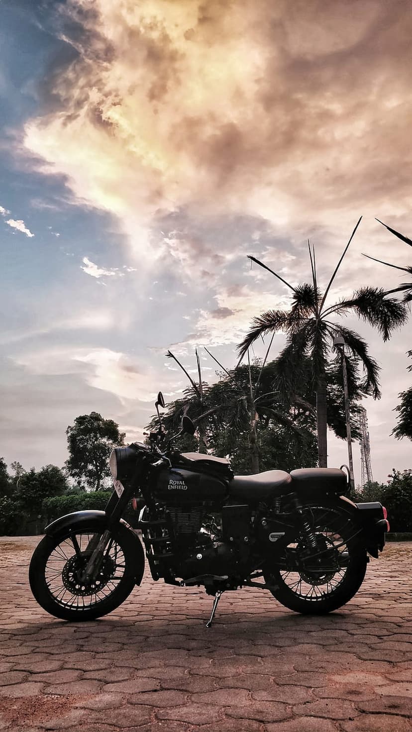 Black Royal Enfield motorcycle parked on bricks against a silhouette of palms and a dramatic sunset sky.
