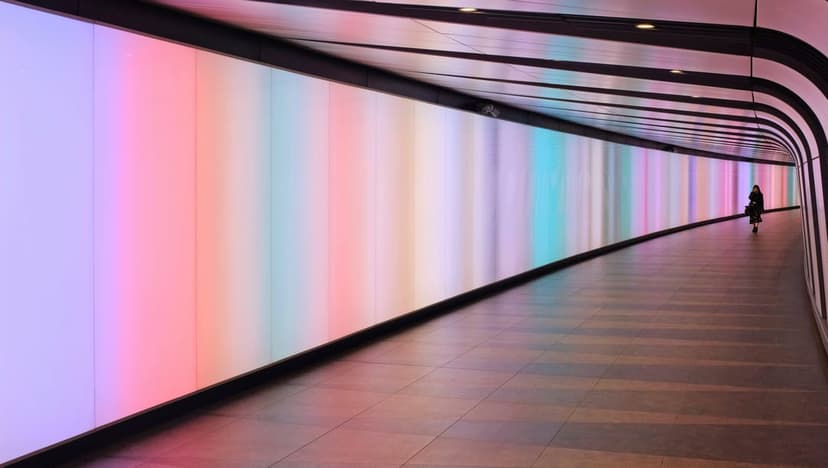 Person walking away through a curved tunnel illuminated by vertical neon light strips in pastel rainbow colors.