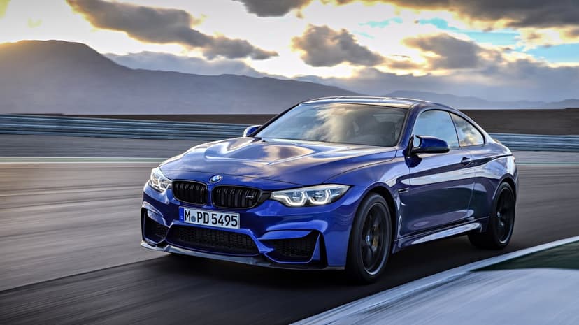 Blue BMW M4 sports car driving on a race track at sunset with mountains and dramatic clouds in the background.