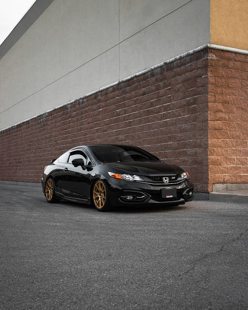 Black Honda Civic coupe with gold wheels parked on asphalt against a brick and cream-colored building wall.
