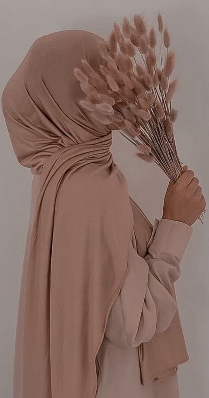 Woman wearing a dusty rose hijab holding a bouquet of dried bunny tail grass against a soft neutral background.