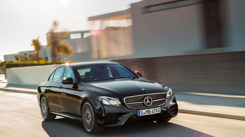 Black Mercedes-Benz sedan driving past modern white buildings with motion blur and warm sunlight lens flare.