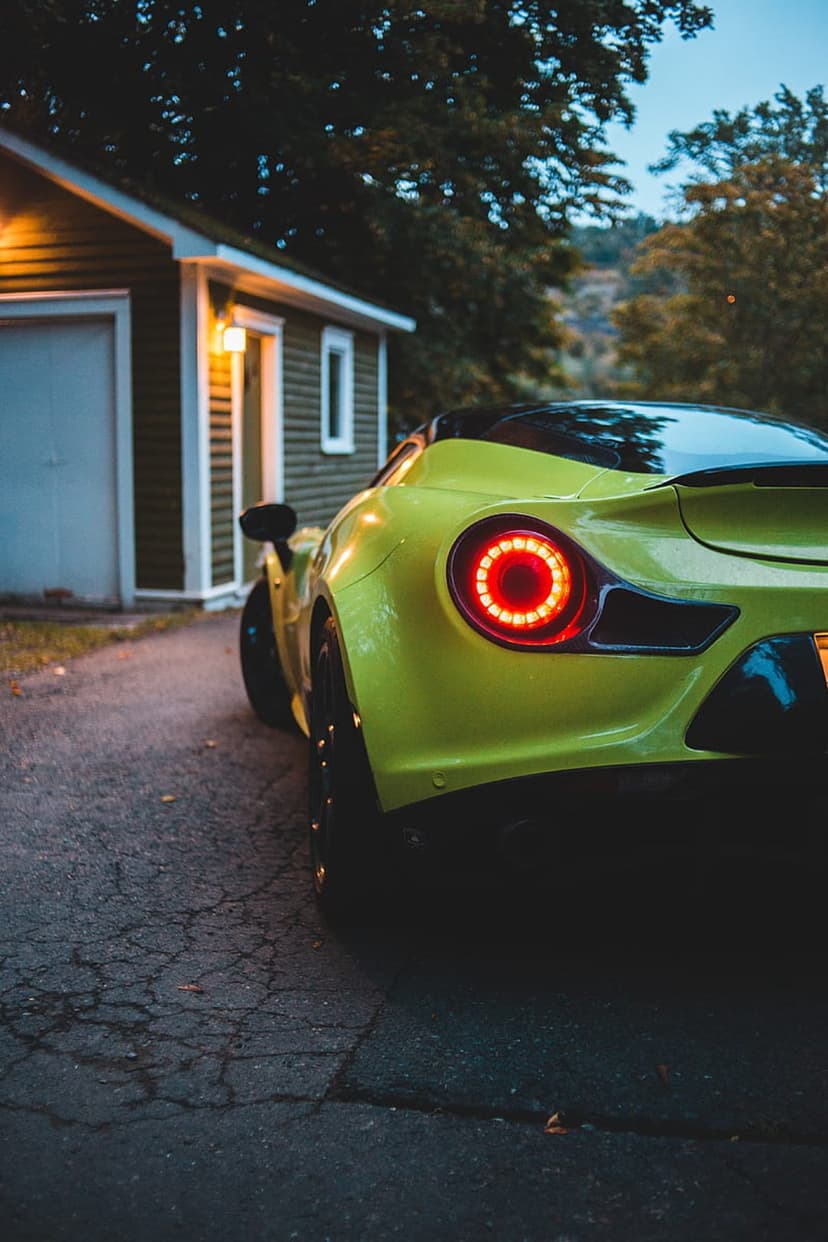 Lime green sports car rear tail light glows red at dusk on a driveway next to a lit garage window and trees.