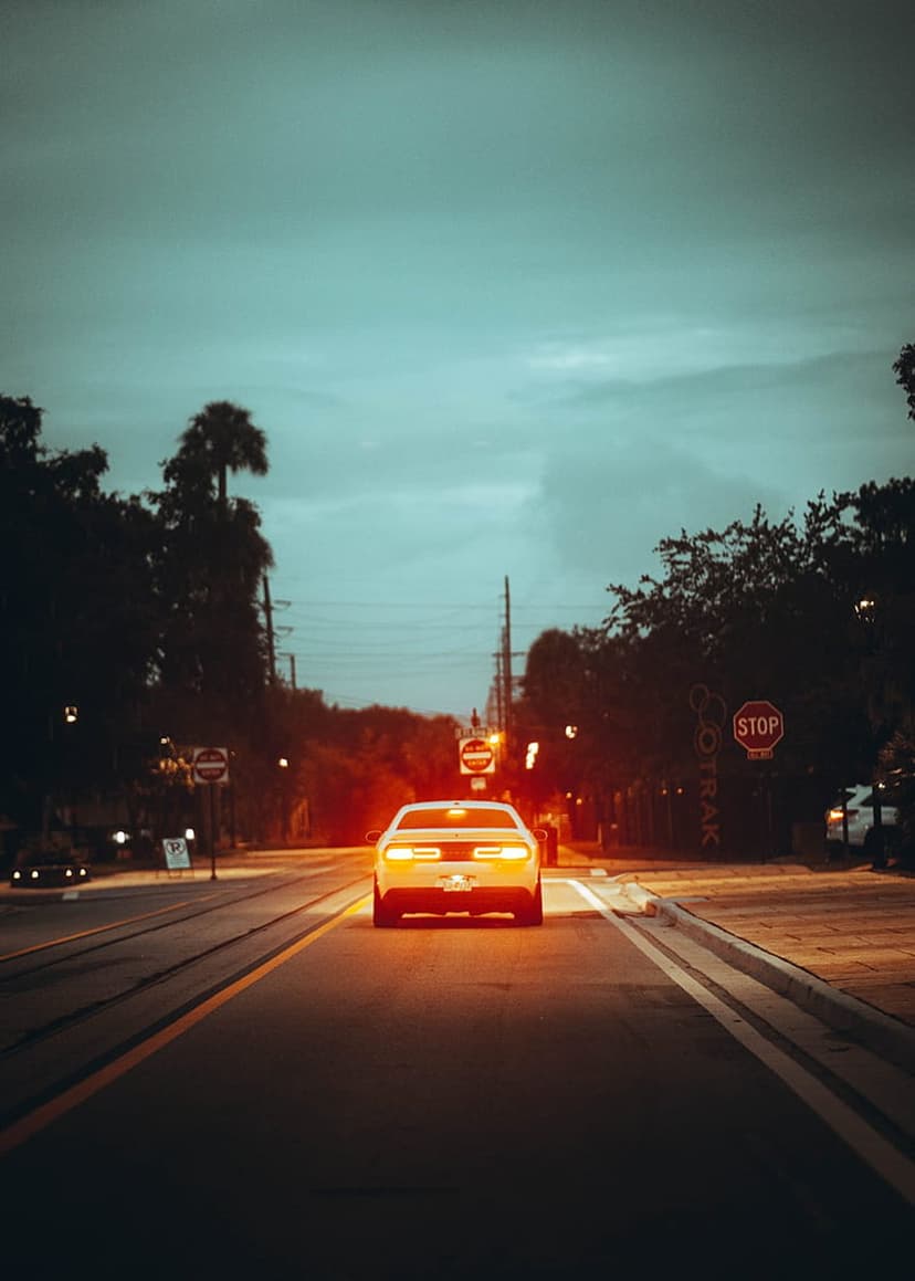 White car driving away on a dark road at dusk with glowing red taillights, stop signs, and teal sky above.