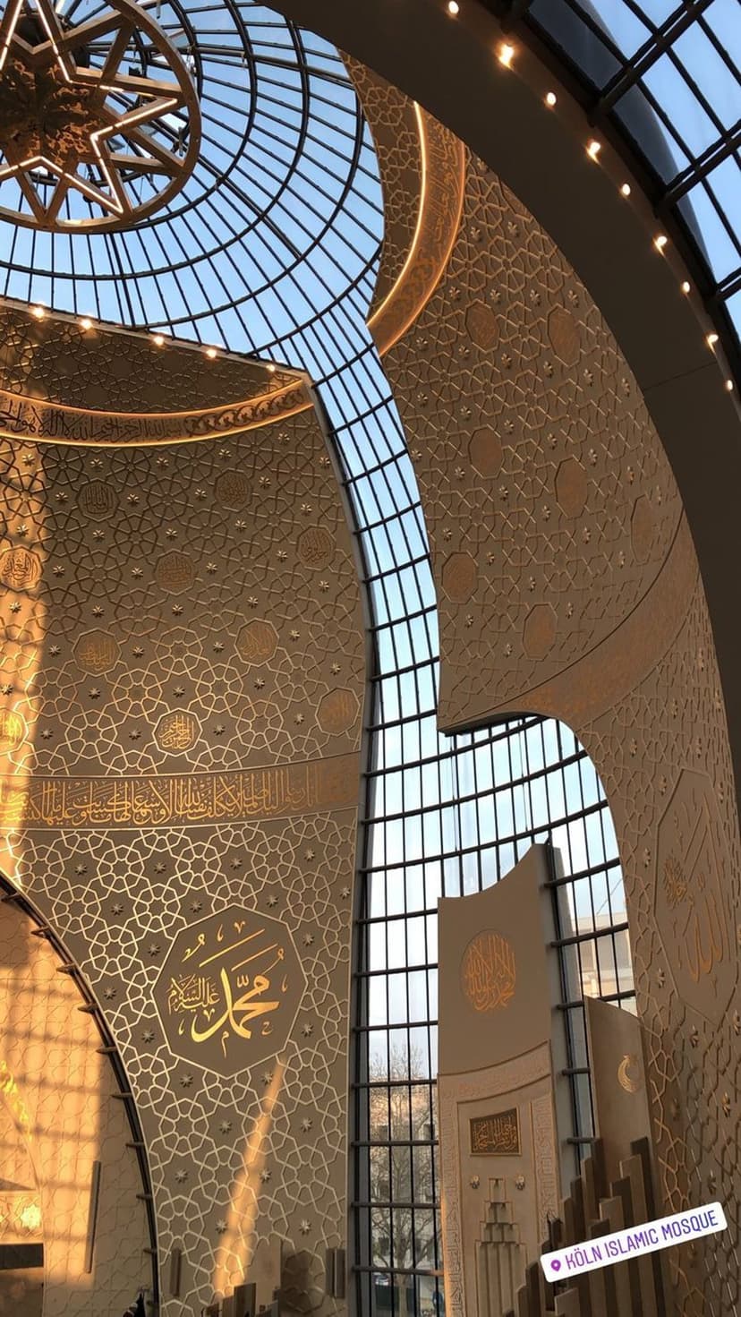 Golden Arabic calligraphy and geometric patterns on a white dome ceiling with sunlight streaming through glass.