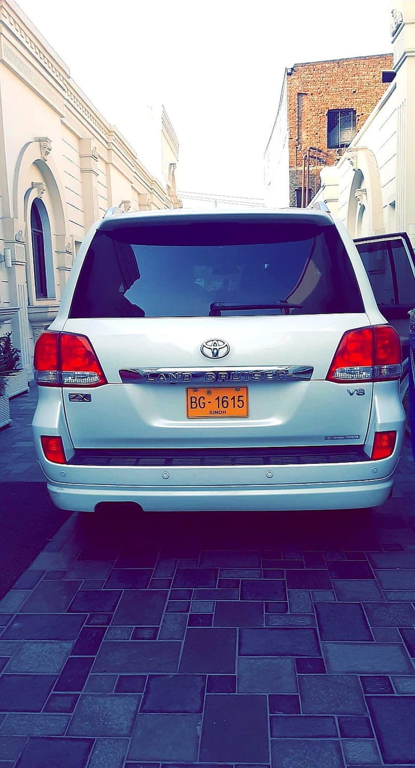 White Toyota Land Cruiser SUV parked on a tiled driveway between buildings with a yellow license plate visible.