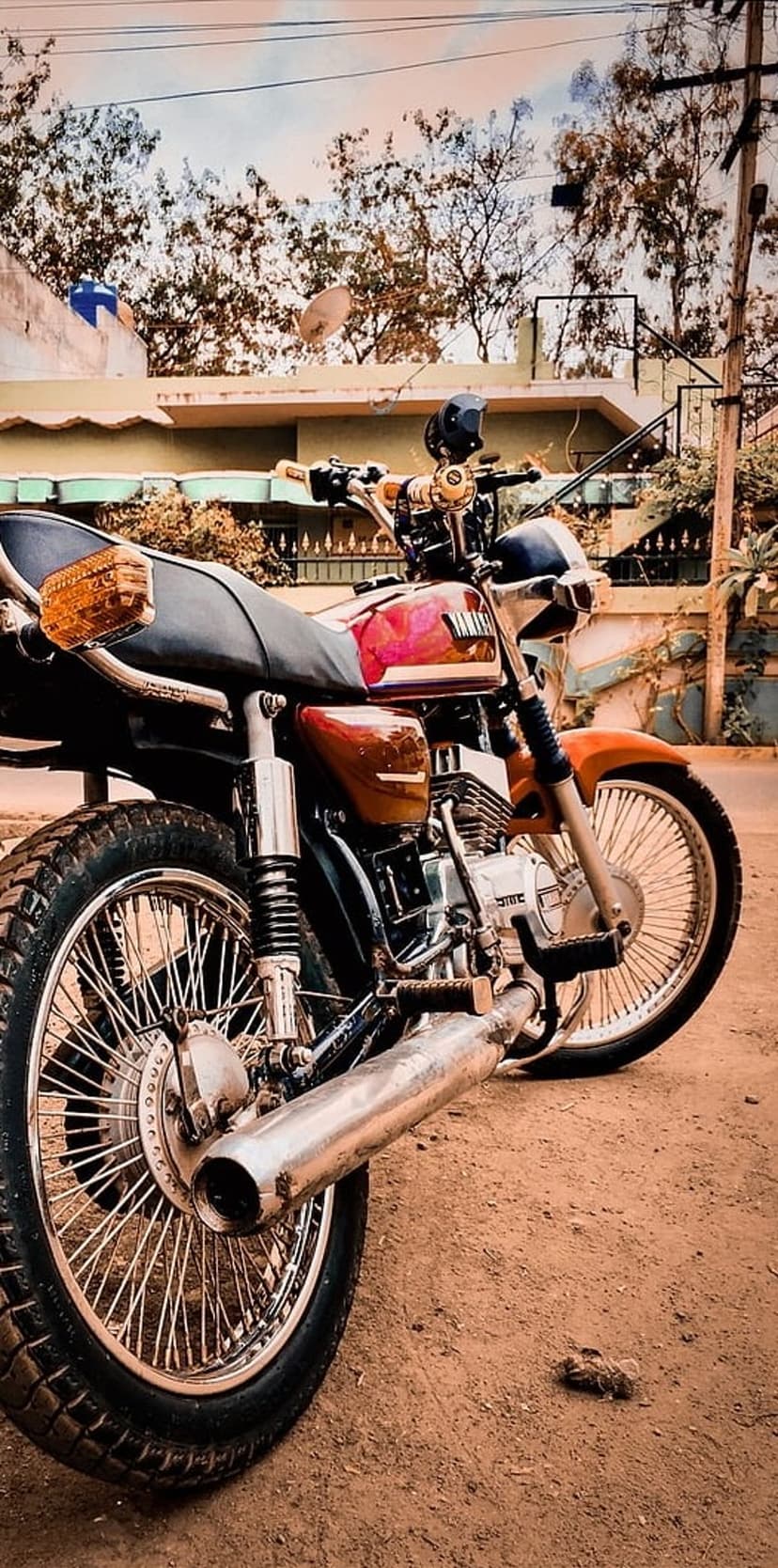 Red vintage Yamaha RX 100 motorcycle with chrome exhaust parked on a dusty rural road with green foliage.