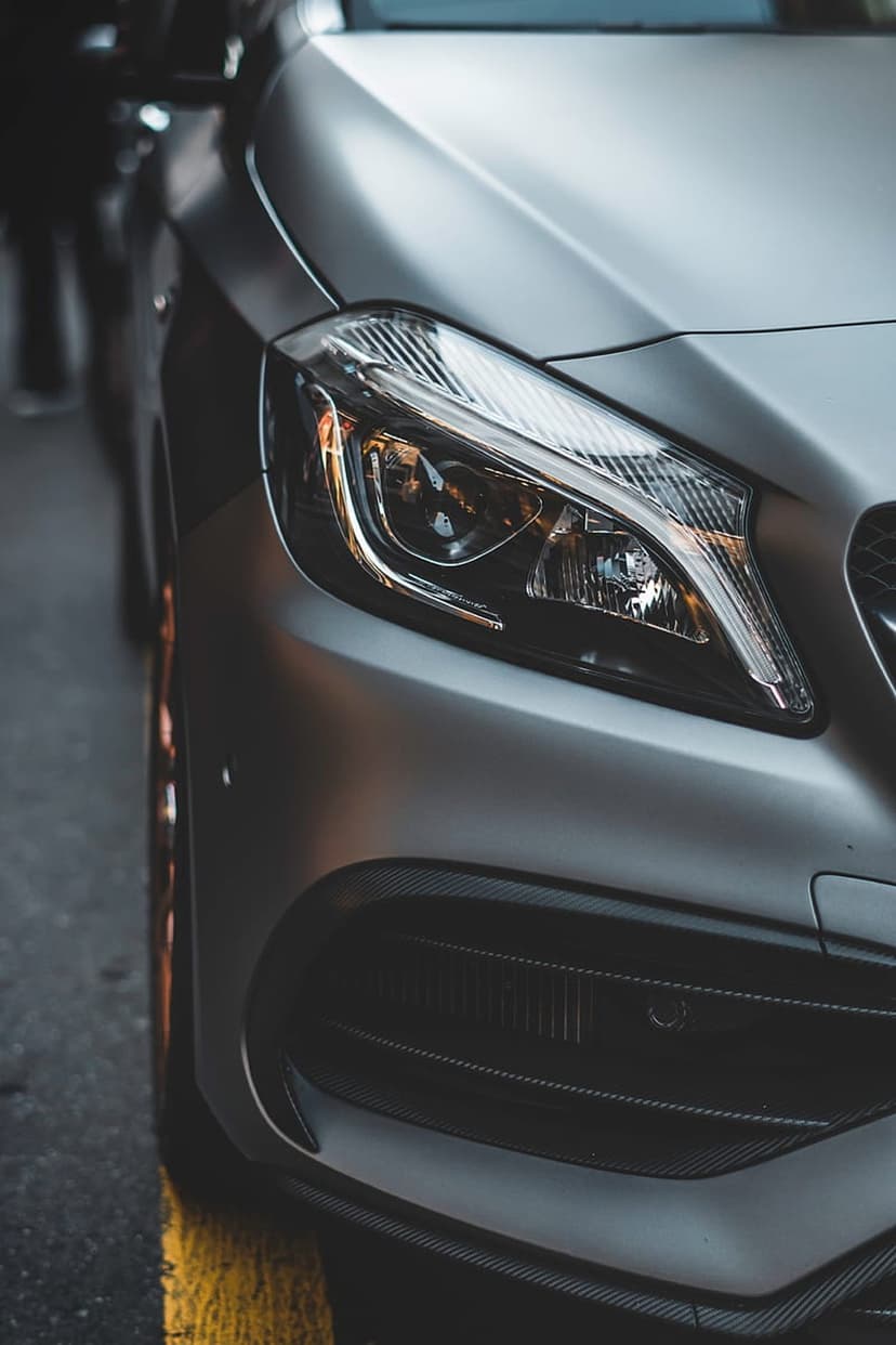 Silver car headlight with LED optics and carbon fiber bumper accents on a wet urban road with yellow markings.