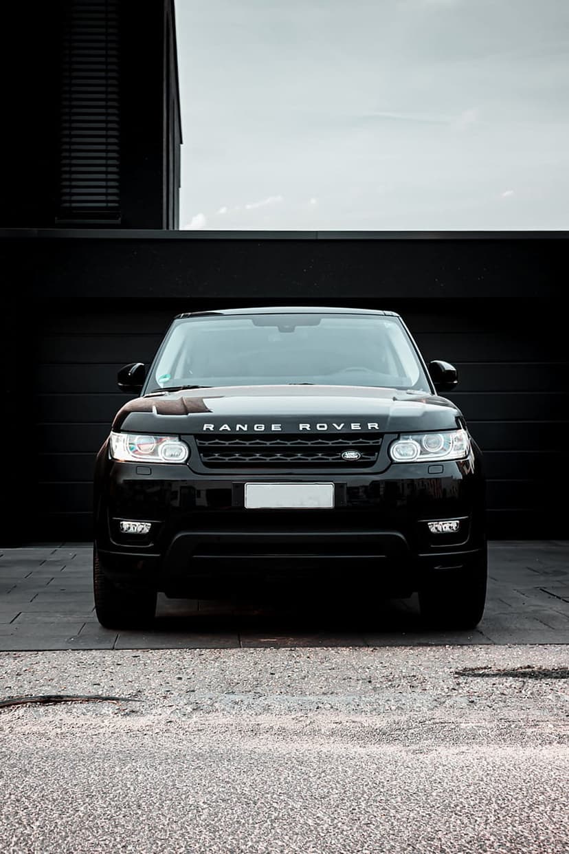 Black Range Rover with lit headlights parked on a gravel driveway in front of a dark contemporary garage.