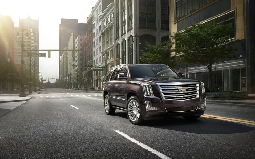 Dark purple Cadillac Escalade on a city street with morning sunlight, chrome grille, and urban buildings.