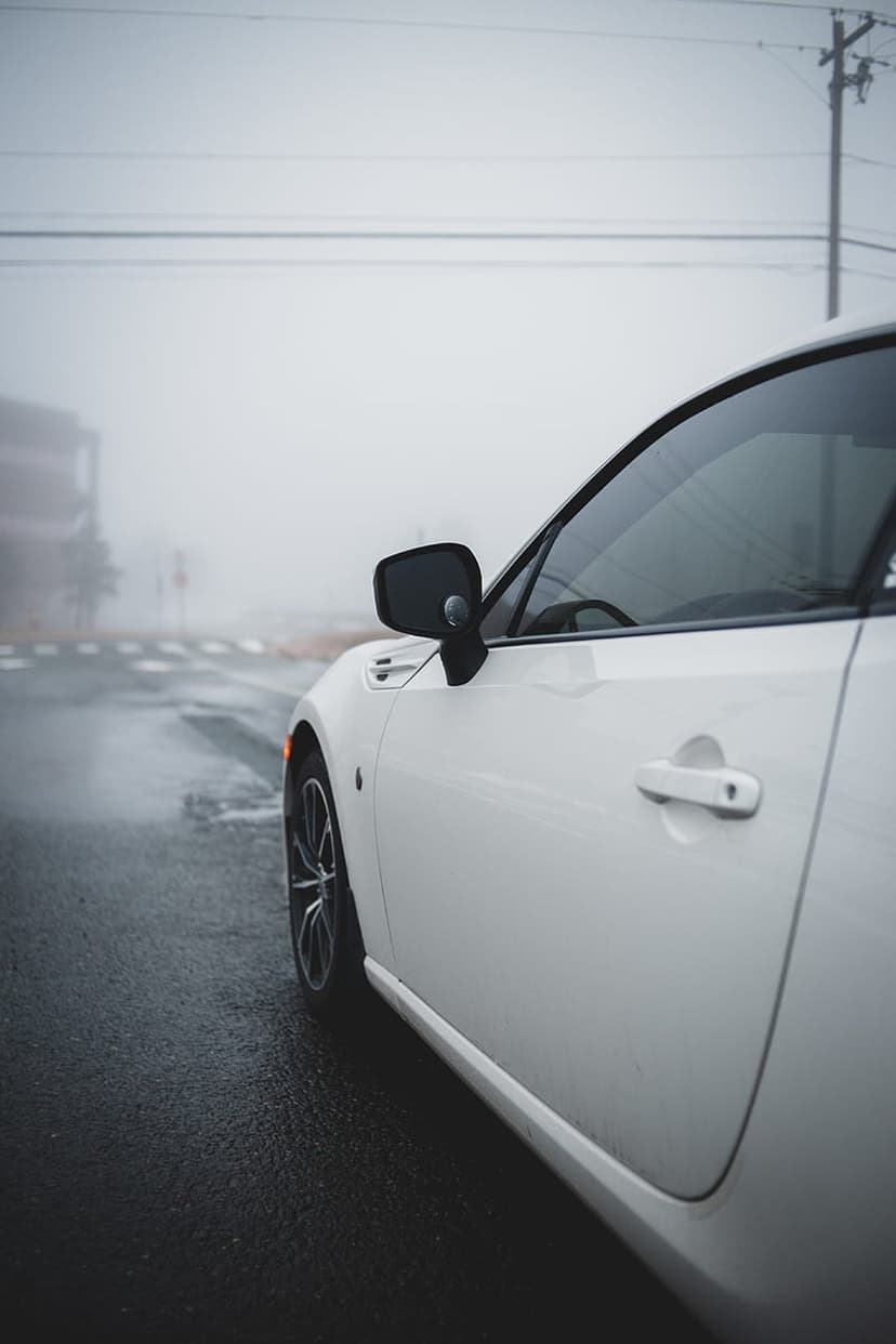 White sports car side profile on wet asphalt during a foggy night with a visible wheel and door handle.