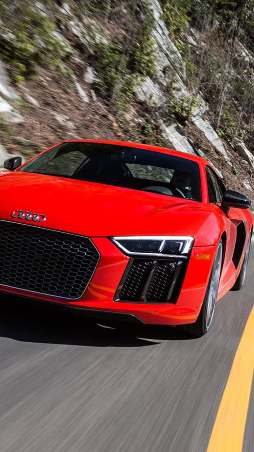 Red Audi R8 sports car maneuvers on a mountain road with aggressive LED headlights and motion-blurred scenery.