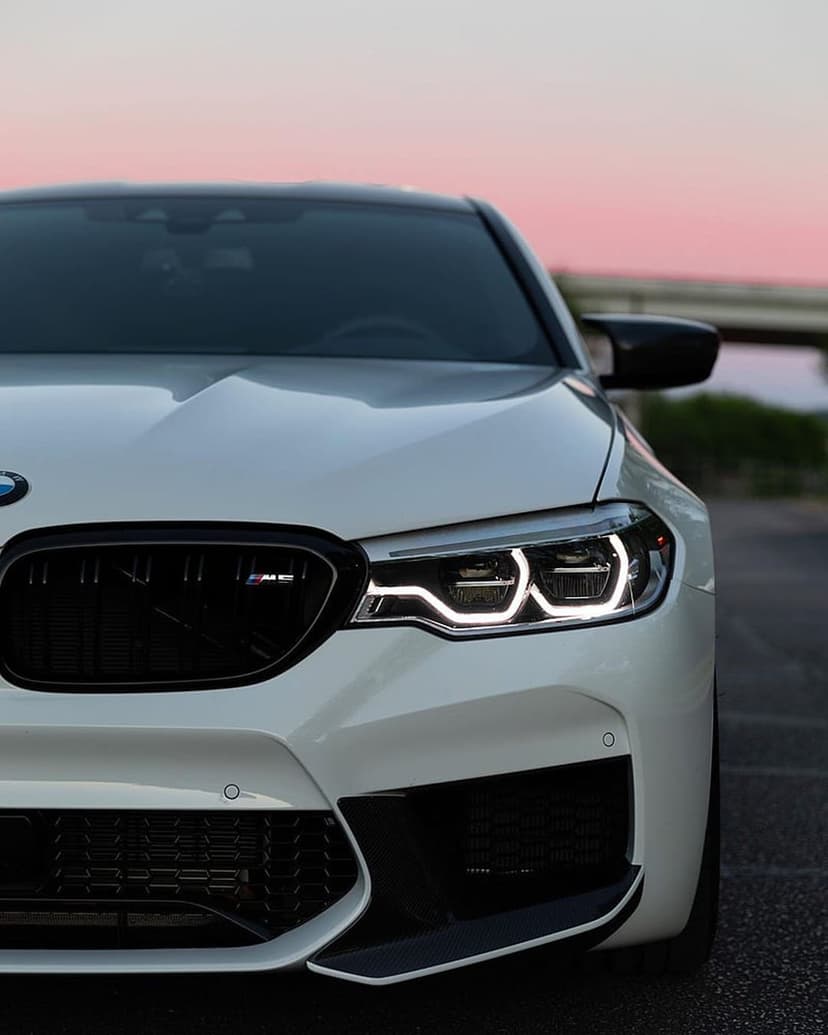 BMW M5 LED headlights and kidney grille glowing under a soft sunset sky on a clean white sports sedan exterior.