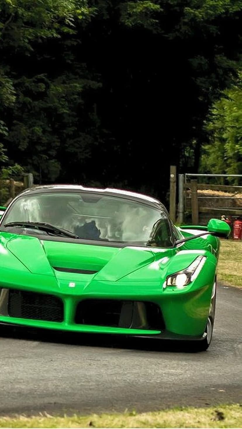 Bright green Ferrari supercar drives along a winding road lined with lush trees and a rustic wooden fence.
