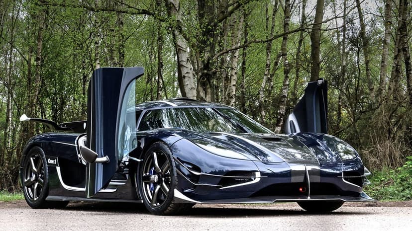 Dark blue Koenigsegg One:1 with open doors parked on a gravel road surrounded by green forest trees.