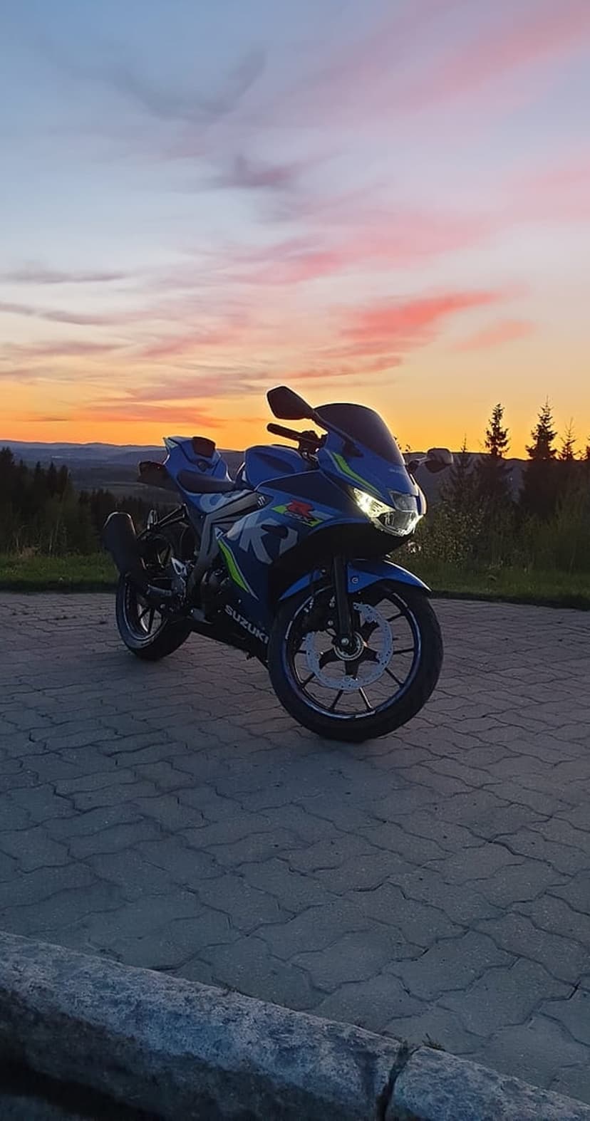 Blue Suzuki sportbike on a stone patio during a vibrant pink and orange sunset with a scenic twilight sky.
