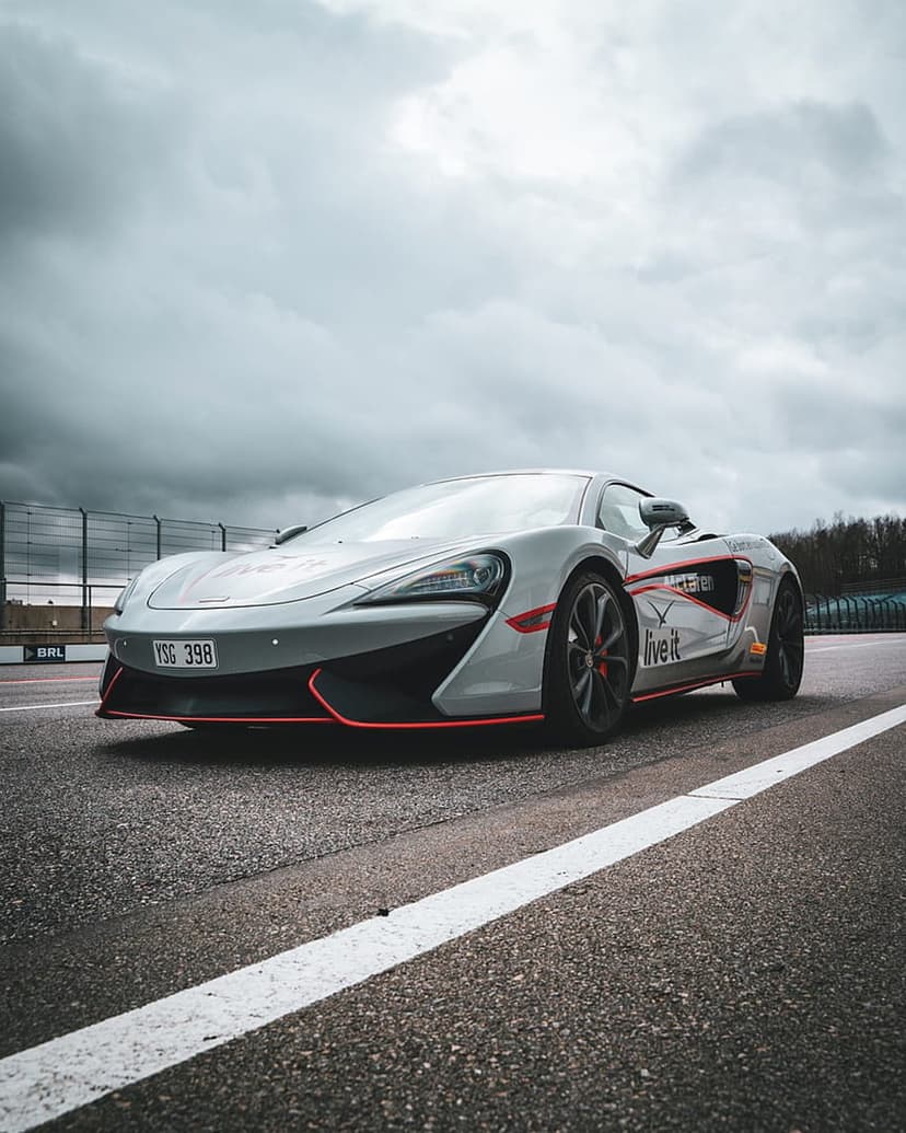 McLaren 570S supercar in silver with red accents parked on a race track under a dark, dramatic overcast sky.