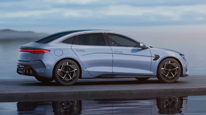Sleek light blue electric sedan parked on a concrete pier with water reflections under a soft cloudy sky.