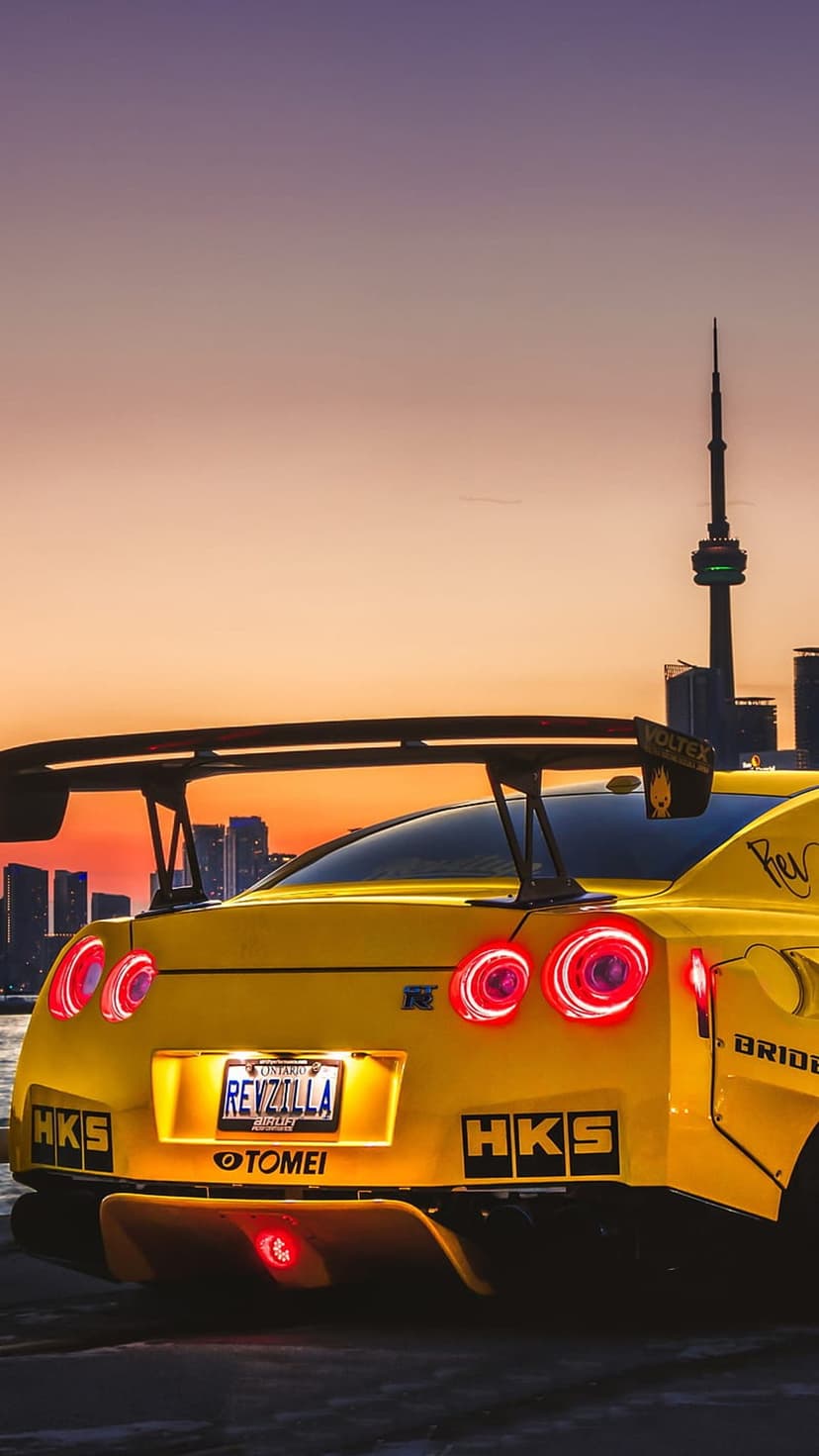 Yellow Nissan GT-R rear view with glowing red taillights against a sunset city skyline and tall tower.