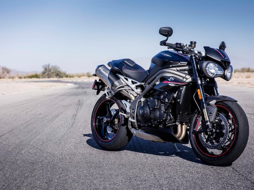 Black Triumph motorcycle with dual headlights parked on a gray asphalt racetrack under a clear blue sky.