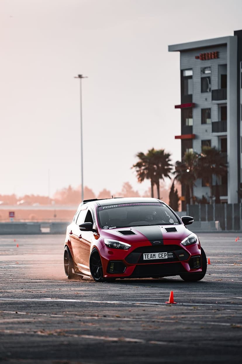 Red Ford Focus RS drifts on a track, kicking up gray smoke near palm trees and orange cones under a hazy sky.
