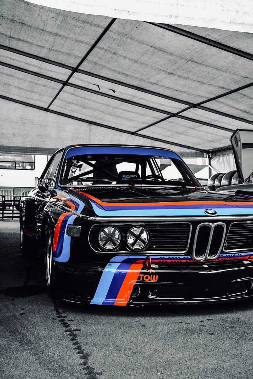 Black BMW E46 M3 race car with blue and red stripes, front splitter, and rally headlights under a canopy.