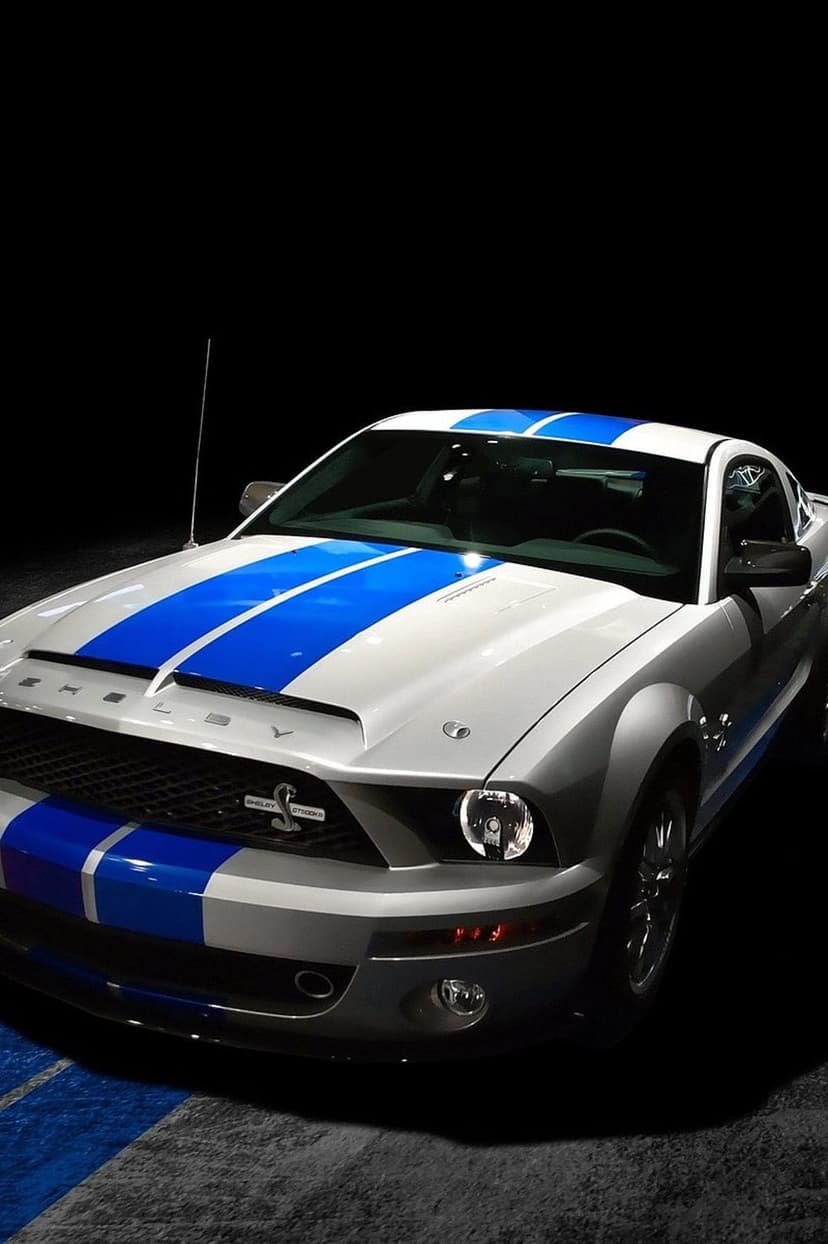 White Shelby Mustang with blue racing stripes, aggressive front grille, and cobra badge on a dark background.