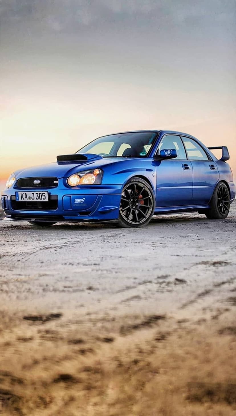Blue Subaru WRX STI parked on a sandy beach during golden hour with its rear spoiler and hood scoop visible.