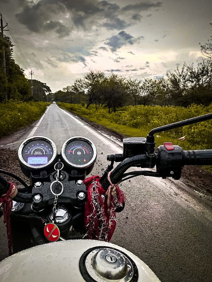 Motorcycle handlebars and dashboard overlook a glistening wet road lined with green trees under a gray sky.