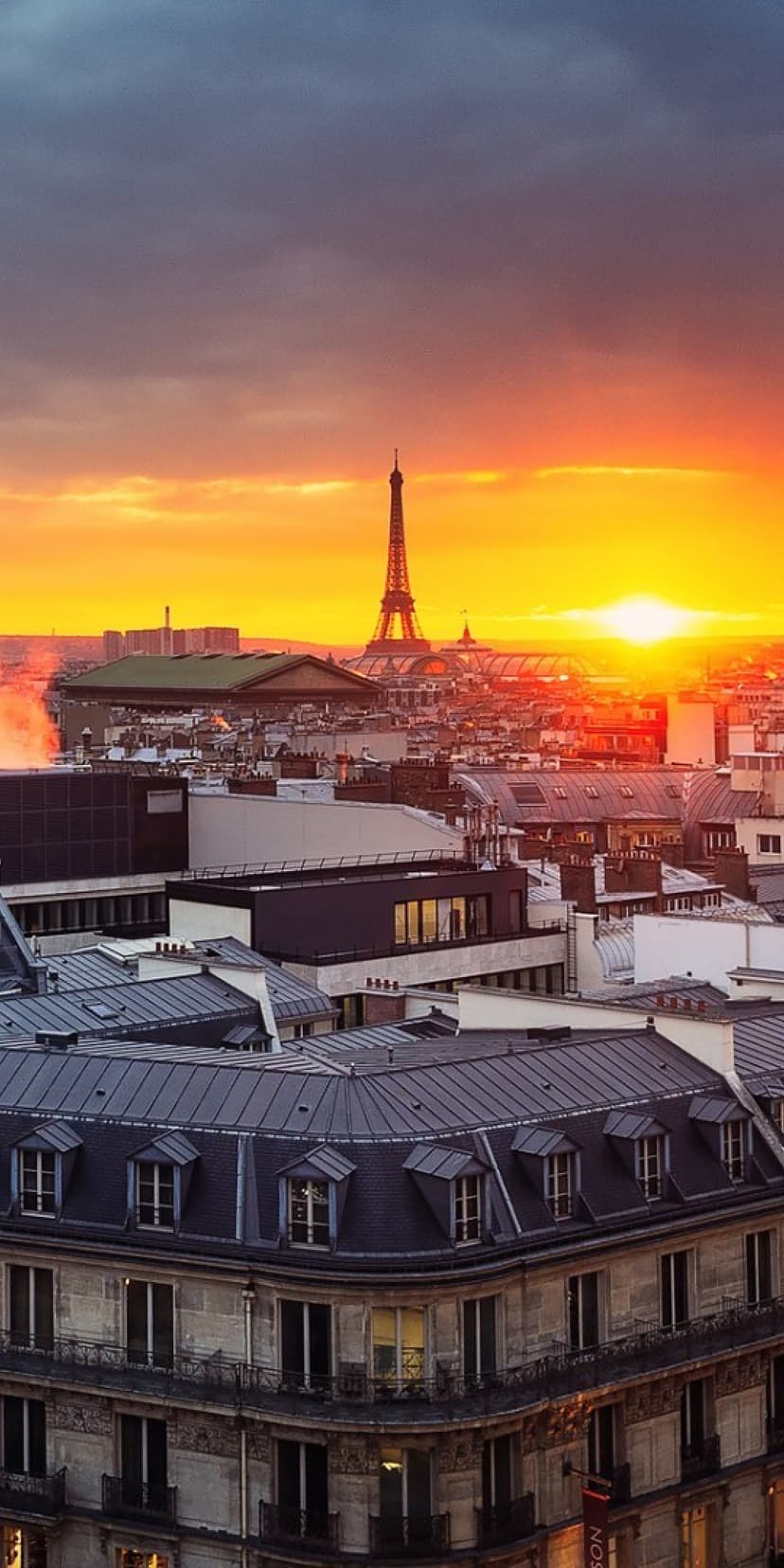 Eiffel Tower silhouetted against an orange sunset sky behind classic Parisian zinc rooftops and chimneys.