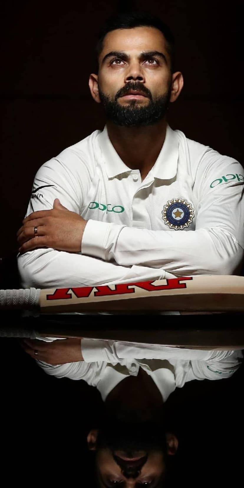 Virat Kohli in white cricket attire looks upward behind the symmetrical reflection of an MRF bat on a dark backdrop.