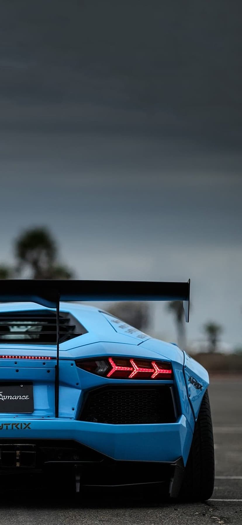 Blue Lamborghini Aventador rear view featuring a large black wing, red taillights, and a cloudy sky background.